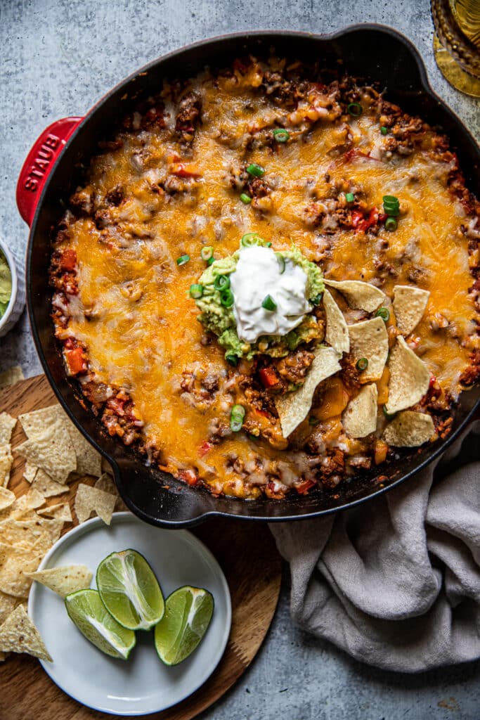 A cast iron skillet with feef taco filling and melted cheese on top.  It's served with guacamole and sour cream on top.  There are limes and chips off to the side for serving too.  A linen napkin is placed to the other side with more guacamole