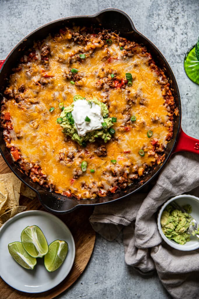 A cast iron skillet with feef taco filling and melted cheese on top.  It's served with guacamole and sour cream on top.  There are limes and chips off to the side for serving too.  A linen napkin is placed to the other side with more guacamole