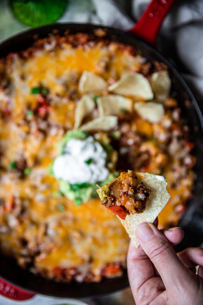 A close up of a cast iron skillet with feef taco filling and melted cheese on top.  It's served with guacamole and sour cream on top.  There's a hand in the foreground with a chip and filling scooped on it.
