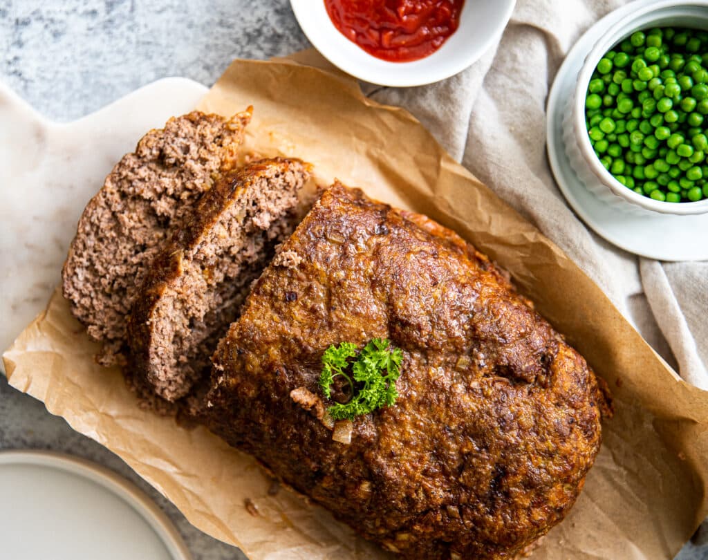 A meatloaf laid out on a marble cutting board over parchment paper.  There is also a bowl of peas to one side and a bowl of ketchup. 