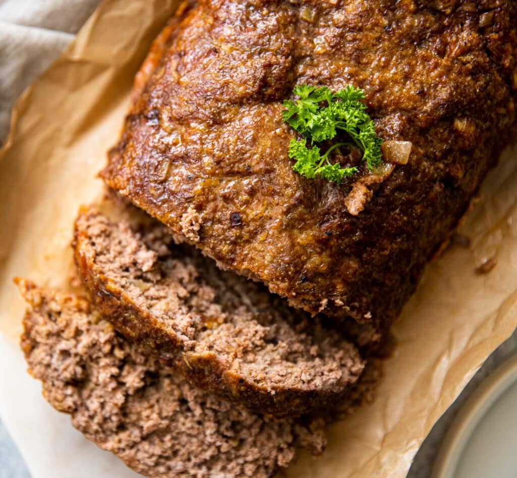 A close up of a meatloaf loaf.  It's set on parchment paper on a cutting board.  there is a small piece of parsley on top.
