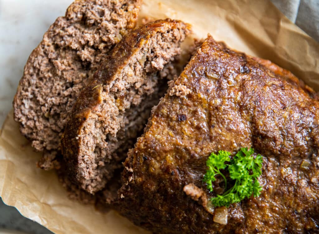 A close up of a meatloaf loaf with three sliced cut.  It's set on parchment paper on a cutting board.  there is a small piece of parsley on top.