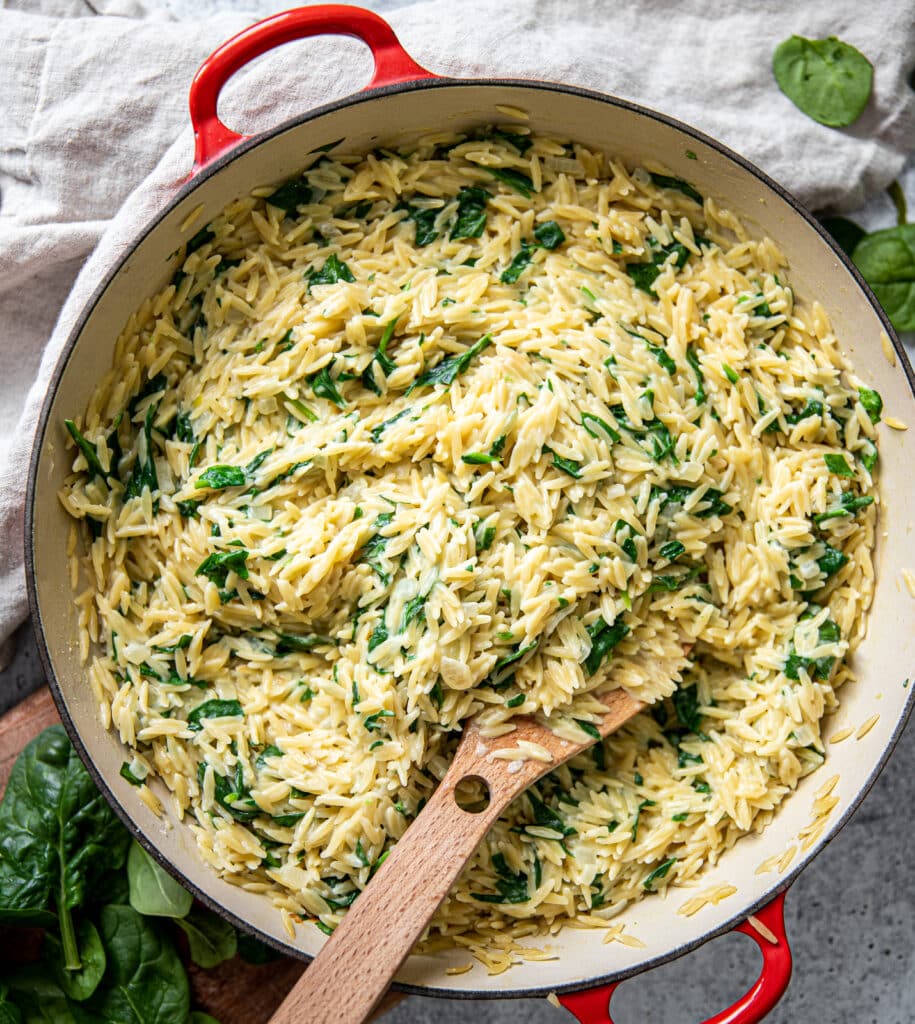 A large red pot with creamy parmesan orzo with lots of spinach.  There is decorative spinach to the top right and bottom left.