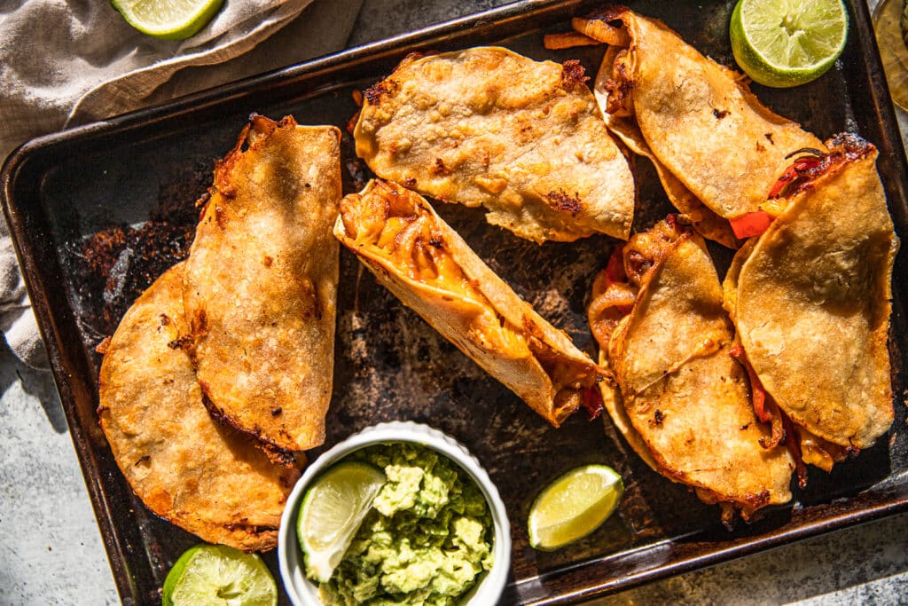 A baking sheet with crispy shrimp and pepper tacos spread out over it.  Guacamole and limes places around baking sheet for extra flavor
