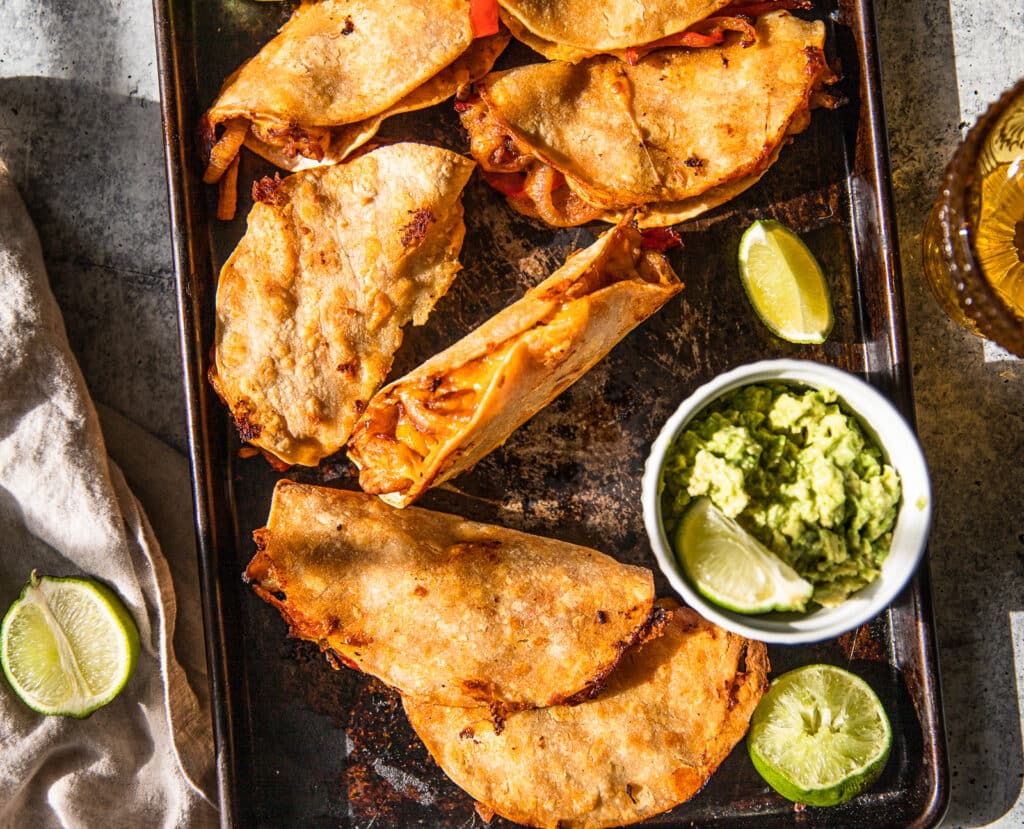 A baking sheet with crispy shrimp and pepper tacos spread out over it.  Guacamole and limes places around baking sheet for extra flavor