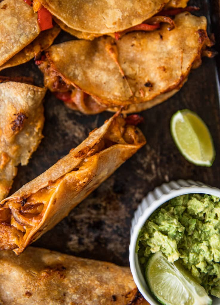 A baking sheet with crispy shrimp, cheese, and pepper tacos spread out over it.  Guacamole and limes places around baking sheet for extra flavor