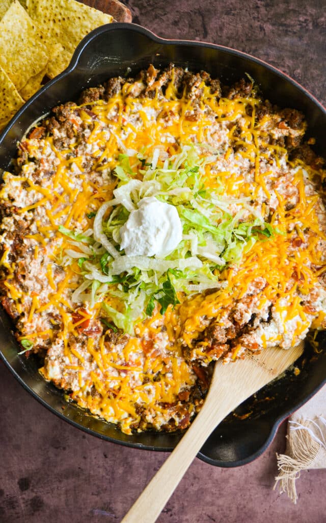 a cast iron skillet on a table.  In the skillet is ground beef, cheese, and salsa topped with shredded lettuce and sour cream.  A wooden spoon is in the skillet to the side for serving.