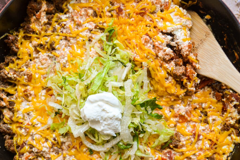 a cast iron skillet on a table.  In the skillet is ground beef, cheese, and salsa topped with shredded lettuce and sour cream.  A wooden spoon is in the skillet to the side for serving.