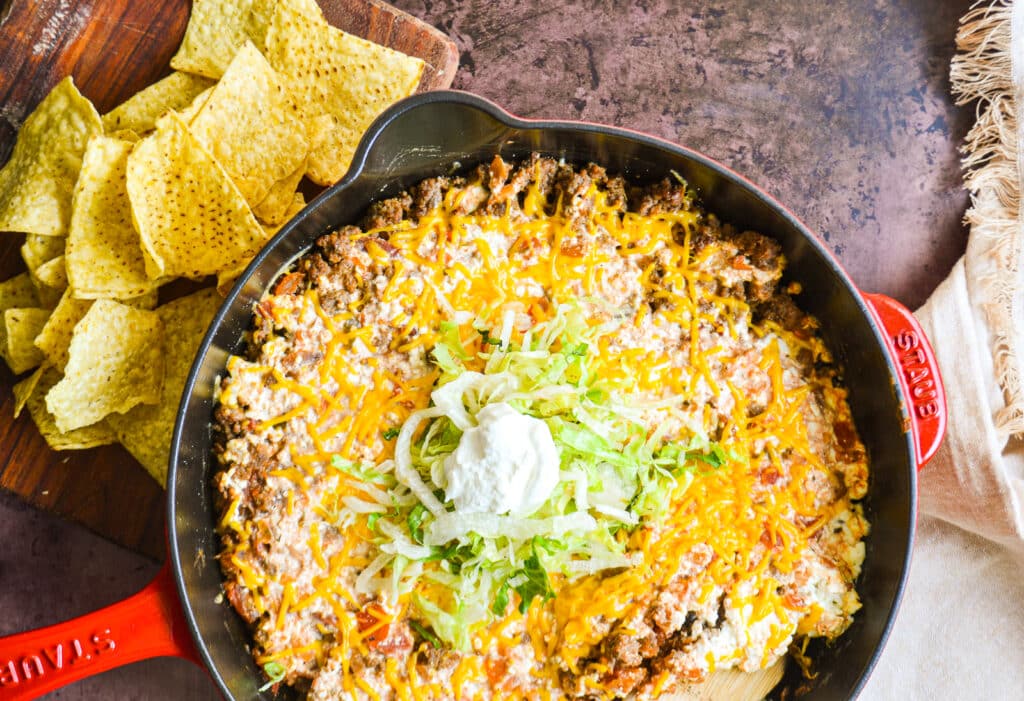 a cast iron skillet on a table.  In the skillet is ground beef, cheese, and salsa topped with shredded lettuce and sour cream.   There are chips to the other side/