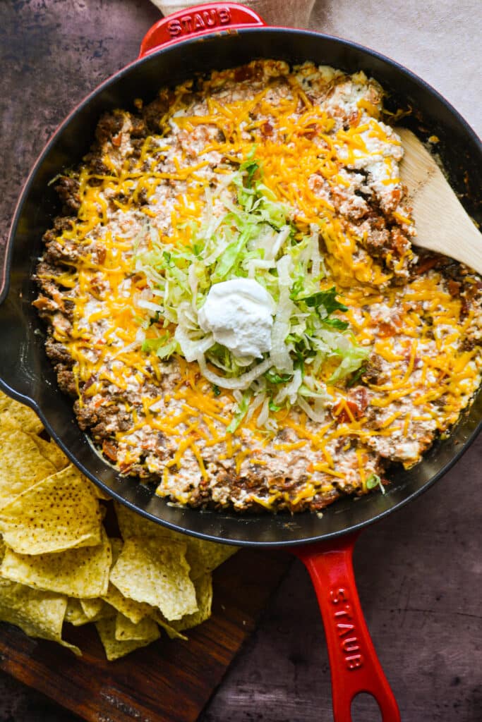 a cast iron skillet on a table.  In the skillet is ground beef, cheese, and salsa topped with shredded lettuce and sour cream.  A wooden spoon is in the skillet to the side for serving.  There are chips to the other side/